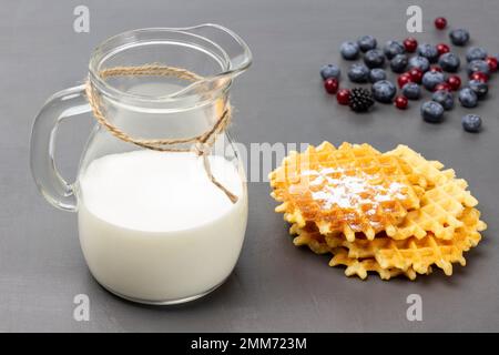 Milk in glass jug. Waffles and berries on table. Top view. Grey background. Stock Photo