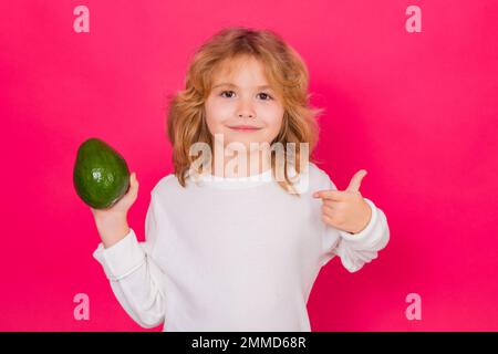 Kid hold red avocado in studio. Studio portrait of cute child with avocado isolated on red background. Stock Photo
