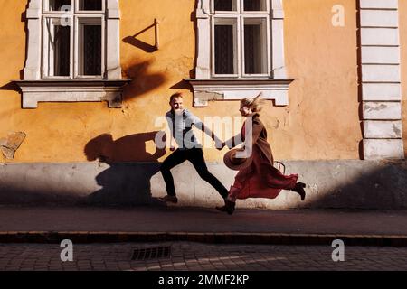 Stylish cheerful young hipsters bearded man and blonde woman are holding hands having fun at old European city streets. Happy couple in love is runnin Stock Photo