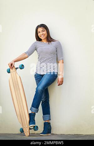 My day isnt complete without a skate. a young skater standing against a wall with her longboard. Stock Photo