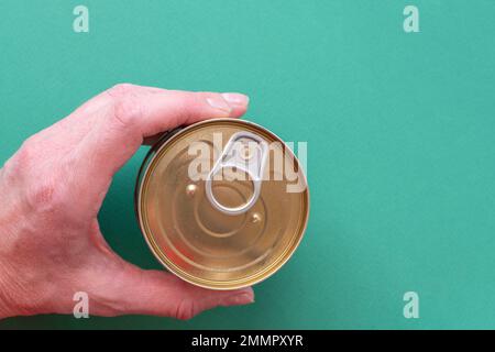 The hand of an adult man holds a closed can of canned food with a public key on a green background. Top view of tin can with ring pull isolated on gre Stock Photo