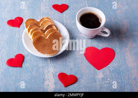 Homemade pancakes in the shape of a heart on a white plate, a mug with coffee or cocoa on a blue stylish wooden background with hearts. Breakfast for Stock Photo