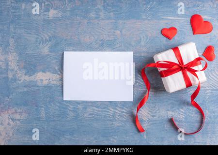 Happy Valentine's Day greetings. Blank white sheet of paper, next to a gift with red hearts on wooden grunge blue background, top view, copy space. Lo Stock Photo