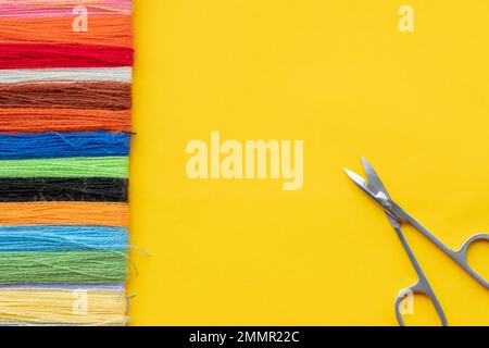 Multi-colored thread for embroidery laid out in a row on a yellow background and scissors for cutting. Top view, copy space, flat lay Stock Photo