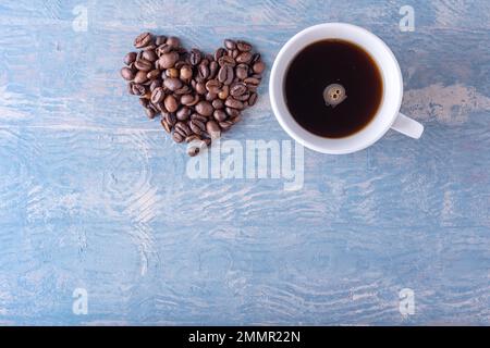 Heart shape made from coffee beans and white coffee cup on blue stylish wooden background, close-up, top view, flat lay. Symbol of love for coffee. Stock Photo