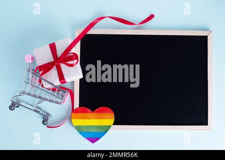 Gift box in shopping carts, rainbow paper heart and blank board with copy space on light blue background. Seasonal sale, discounts, black friday conce Stock Photo