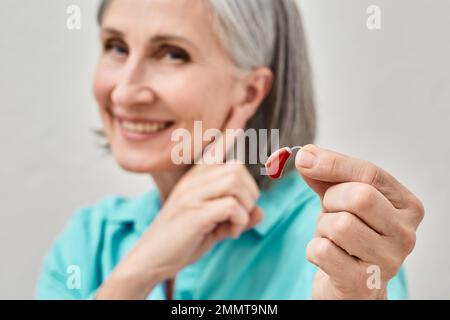 Senior woman holding hearing aid in hand on foreground and pointing finger at her ear, soft focus. Treatment of deafness in people with hearing aids Stock Photo