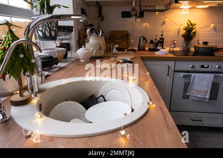 Dirty dishes in the sink in the garlanded kitchen, a holiday after a feast at Christmas. Mess after the guests for the New Year, kitchen cleaning, det Stock Photo