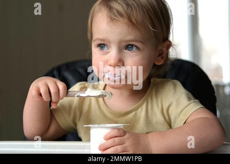 Young Kid Eating Blend Mashed Feed Sitting in High Chair. Baby Weaning. Little Girl Learning to Eat Yogurt, Feeding Himself. Small Hand with Spoon. Br Stock Photo