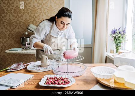 Online classes and workshops on cake decorating and baking. Female Confectioner baker make custom pink heart shape cake in kitchen Bakery. Stock Photo