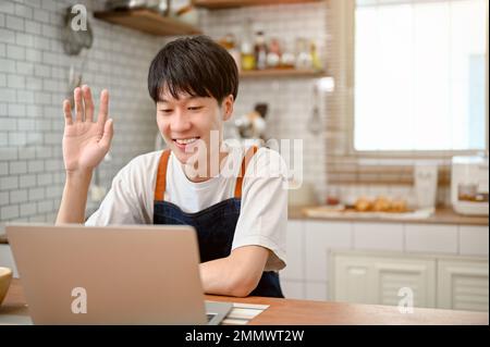 Happy and cute young Asian man in apron using laptop, having an online video call through laptop in the kitchen, waving hand to say hi. Stock Photo