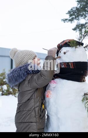 A child paints a snowman's face with paints - winter entertainment and creativity, sculpting a snowman in winter outdoor. Stock Photo