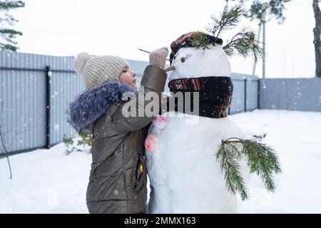 A child paints a snowman's face with paints - winter entertainment and creativity, sculpting a snowman in winter outdoor. Stock Photo