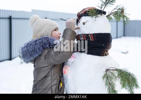 A child paints a snowman's face with paints - winter entertainment and creativity, sculpting a snowman in winter outdoor. Stock Photo