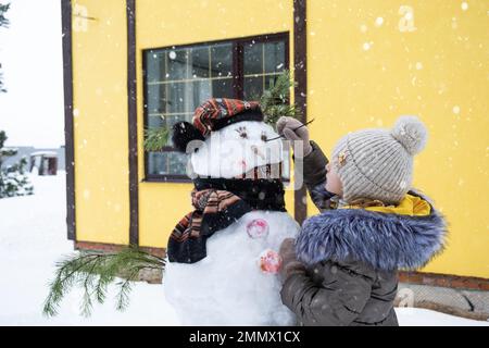 A child paints a snowman's face with paints - winter entertainment and creativity, sculpting a snowman in winter outdoor. Stock Photo