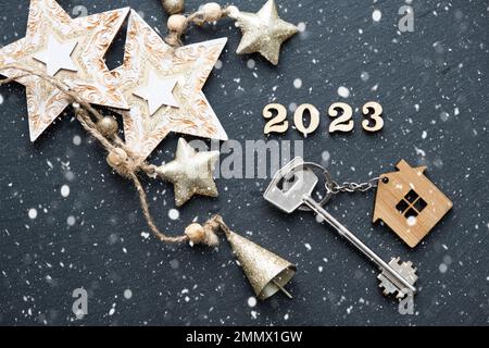 House key with keychain cottage on black background with stars, snowflakes. Happy New Year 2023-wooden letters, greeting card. Purchase, construction, Stock Photo