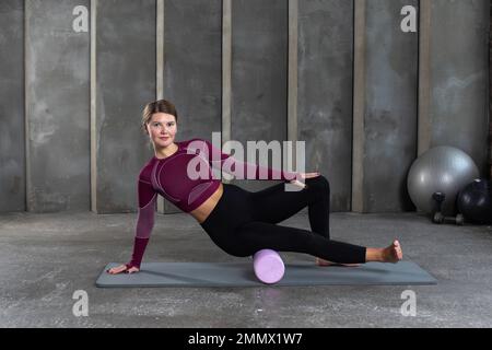 A young woman in sportswear performs a myofascial hip massage with a roller. Muscle recovery, mfr. Stock Photo