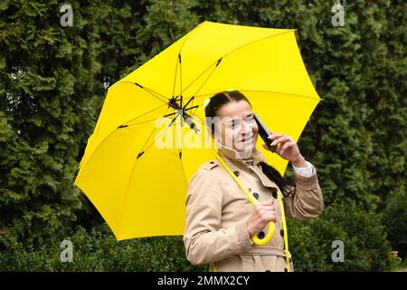 Happy senior woman in yellow rain coat with yellow umbrella is talking on a smartphone and walking in park. Staying in touch with distant loved ones Stock Photo