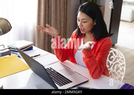 Image of biracial woman sitting at desk talking and gesturing during video call on laptop at home. Working from home, technology, communication and li Stock Photo