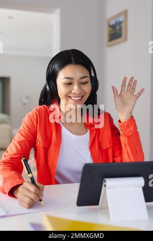 Vertical of happy biracial woman in headphones waving, making tablet video call at home, copy space. Working from home, technology, communication and Stock Photo