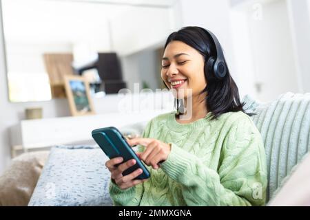 Image of happy biracial woman in headphones using smartphone sitting on sofa at home, copy space. Leisure, relaxation, technology and domestic life co Stock Photo
