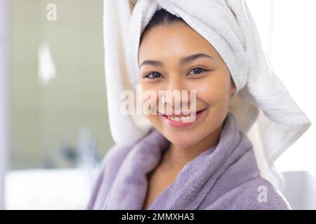 Portrait of happy biracial woman wearing bathrobe and towel smiling in bathroom, with copy space. Relaxation, self care, beauty, leisure and lifestyle Stock Photo