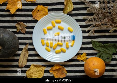 an electronic thermometer lies next to yellow pills on a plate on the table in the kitchen with leaves and pumpkin nearby, medicine and pharmacy, heal Stock Photo
