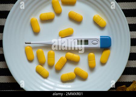 an electronic thermometer lies next to yellow pills on a plate on the table in the kitchen, medicine and pharmacy, health and treatment, pills and ant Stock Photo