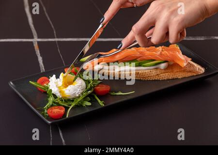 Woman hands cutting poached egg, toast with salmon and avocado on the black background. Stock Photo