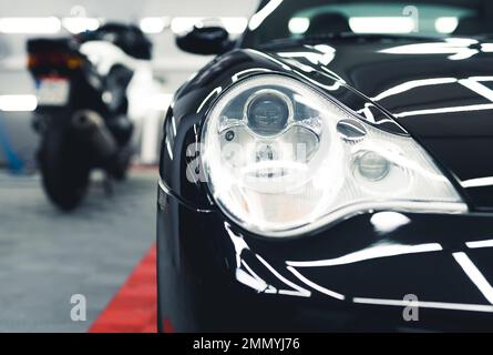 Sports car headlamp. Shiny black sports car in garage. Camera focus on the foreground. Blurred white motorcycle in the background. High quality photo Stock Photo