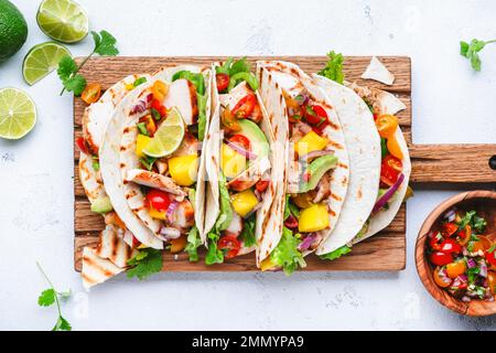 Tacos with grilled chicken, tomato salsa sauce, mango, cilantro and onion in tortilla on cutting board. White table background, top view Stock Photo