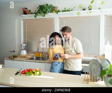 Young heterosexual diverse couple looking at mobile in a kitchen Stock Photo