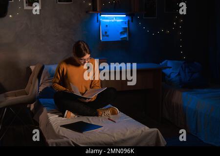 Female university student with long hair sitting on bed doing homework using copybooks and making notes preparing for exam at night Stock Photo