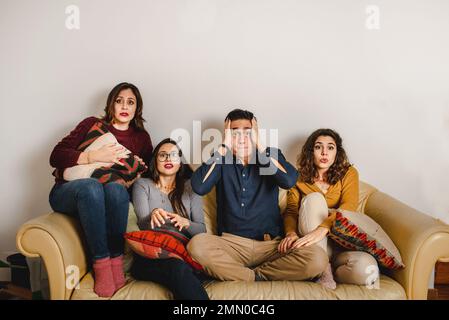 Group of scared friends gathering on sofa and watching horror movie during weekend at home Stock Photo