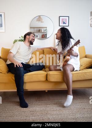 Young diverse heterosexual couple playing guitar and singing on the sofa in an apartment Stock Photo