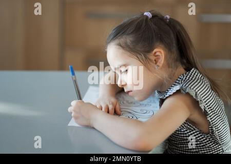 A girl of European appearance in a plaid dress is doing homework for the school at the table in the kitchen. The student thought about the example. The child draws with a pen on paper. High quality FullHD footage Stock Photo