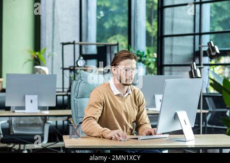 Serious and scorn-centered man in the office at work with a computer, man typing thoughtfully on keyboards, businessman young blond in sweater indoors loft, with green colors, programmer at work. Stock Photo