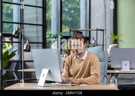 Serious and scorn-centered man in the office at work with a computer, man typing thoughtfully on keyboards, businessman young blond in sweater indoors loft, with green colors, programmer at work. Stock Photo
