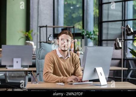 Portrait of thinking serious man with computer inside modern green loft office, blond man looking thoughtfully at camera businessman in casual clothes working with computer. Stock Photo