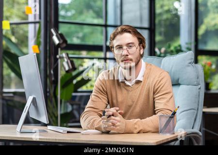 Portrait of thinking serious man with computer inside modern green loft office, blond man looking thoughtfully at camera businessman in casual clothes working with computer. Stock Photo