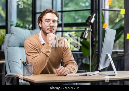 Portrait of thinking serious man with computer inside modern green loft office, blond man looking thoughtfully at camera businessman in casual clothes working with computer. Stock Photo