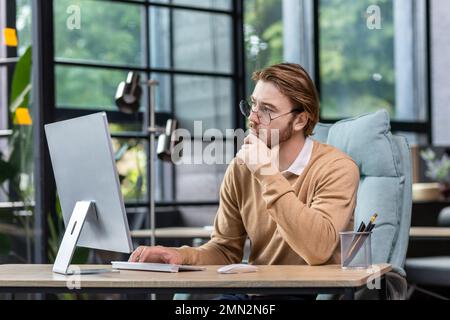 Thinking blond businessman at work inside modern green loft office, young man in casual clothes seriously ponders decision and completes task. Stock Photo