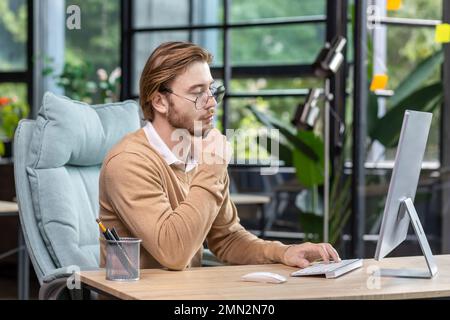 Thinking blond businessman at work inside modern green loft office, young man in casual clothes seriously ponders decision and completes task. Stock Photo