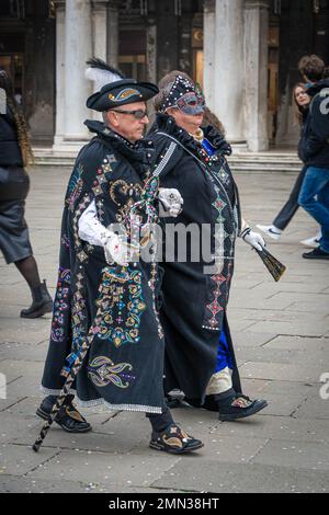 A senior couple dressed in black embroidered carnival mantles walks down the street during Venetian carnival Stock Photo