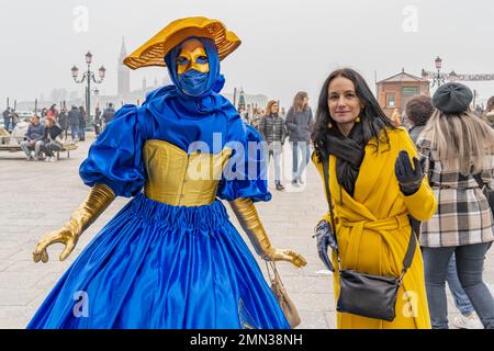 A female in yellow and blue carnaval costume with a flat yellow hat on her hear, wearing a carnival mask, is taking pictures with a female tourist Stock Photo