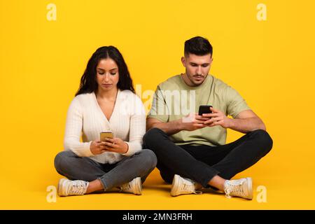 Serious young arab couple in casual sit on floor, chatting and texting on smartphones Stock Photo