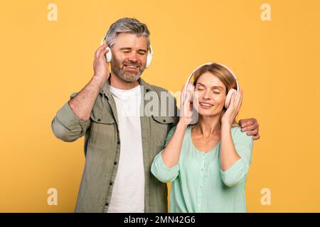 Happy middle aged caucasian spouses wearing headphones and listening to music with closed eyes, yellow background Stock Photo