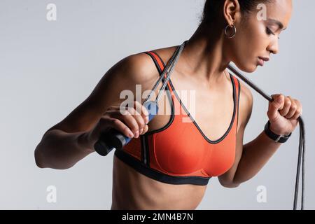 young african american sportswoman in red sports bra holding skipping rope isolated on grey Stock Photo