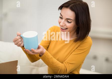 Excited young woman holding blue mug, home interior Stock Photo