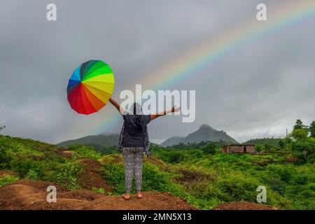 Woman holding colourful umbrella on a trek to Badi Lake in Udaipur India during monsoon rainy season with rainbow in the background Stock Photo